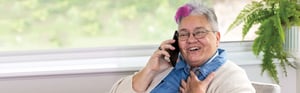 An older woman with a swath of magenta in her gray hair speaks on the phone while using a visible tracheostomy tube.