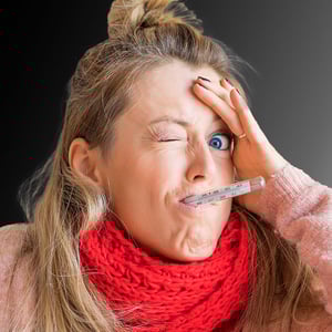 A woman with a thermometer in her mouth holds her head and looks at the camera through one eye