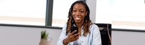 A young Black woman with long hair smiles at the camera as she holds her phone in an office environment.